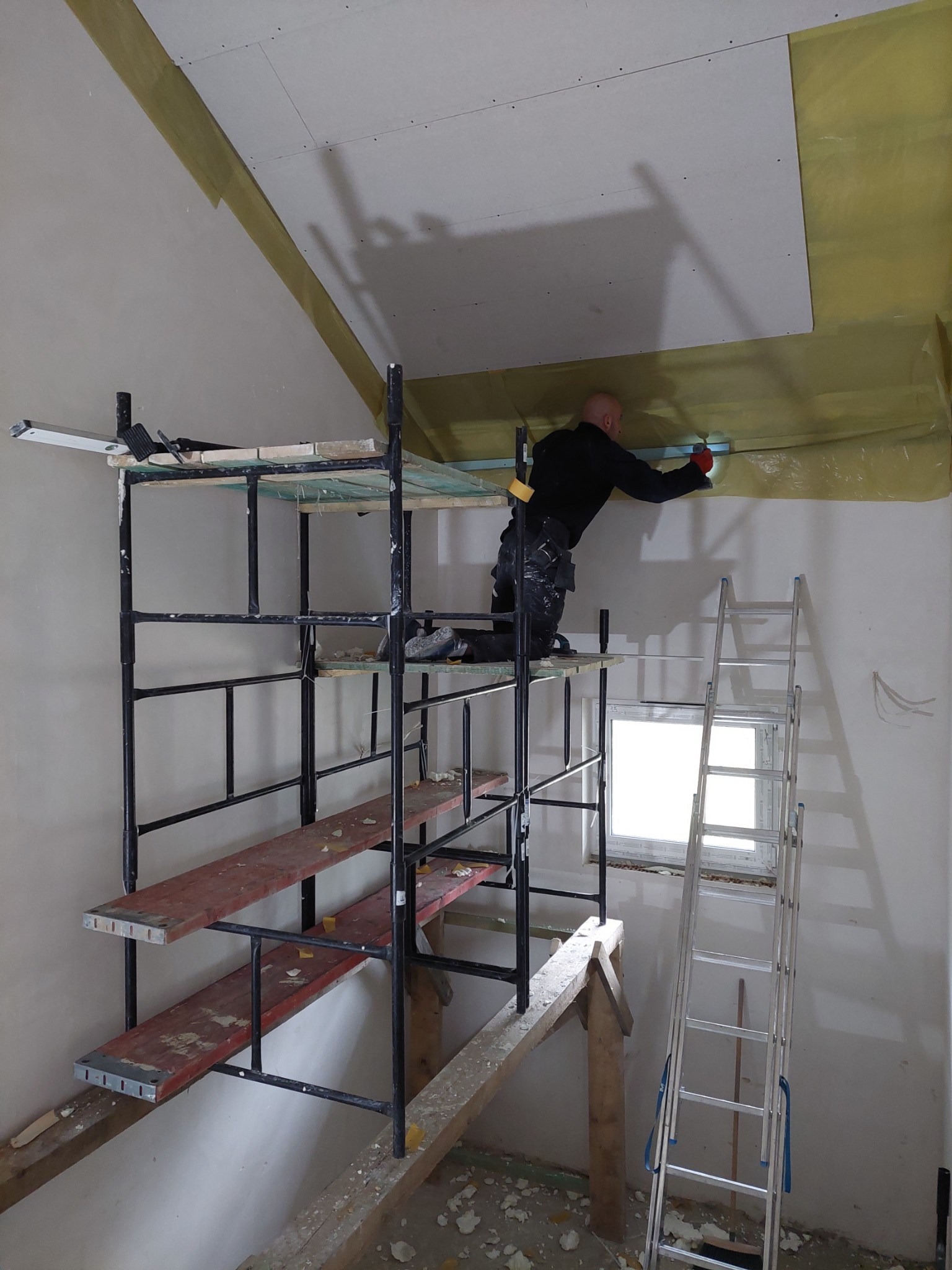 Pracownik w czarnym ubraniu montuje płytę karton gips na suficie poddasza, stojąc na rusztowaniu. Widoczne izolacja z folii żółtej, drabina aluminiowa, okno i fragment ściany. Na rusztowaniu leżą...