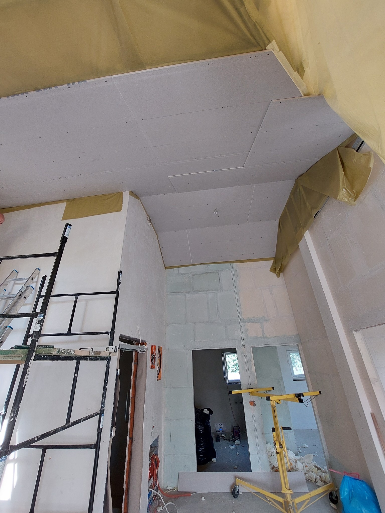 Montaż płyt kartonowo-gipsowych na suficie w Krakowie, widoczny podnośnik do płyt, rusztowanie i zabezpieczenia folią malarską.