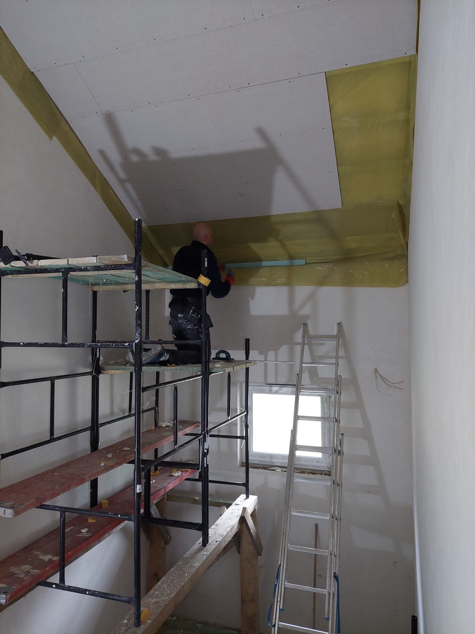 Pracownik na rusztowaniu montuje płytę karton gips na skośnym suficie poddasza, widoczna izolacja z folii. W tle okno i drabina.