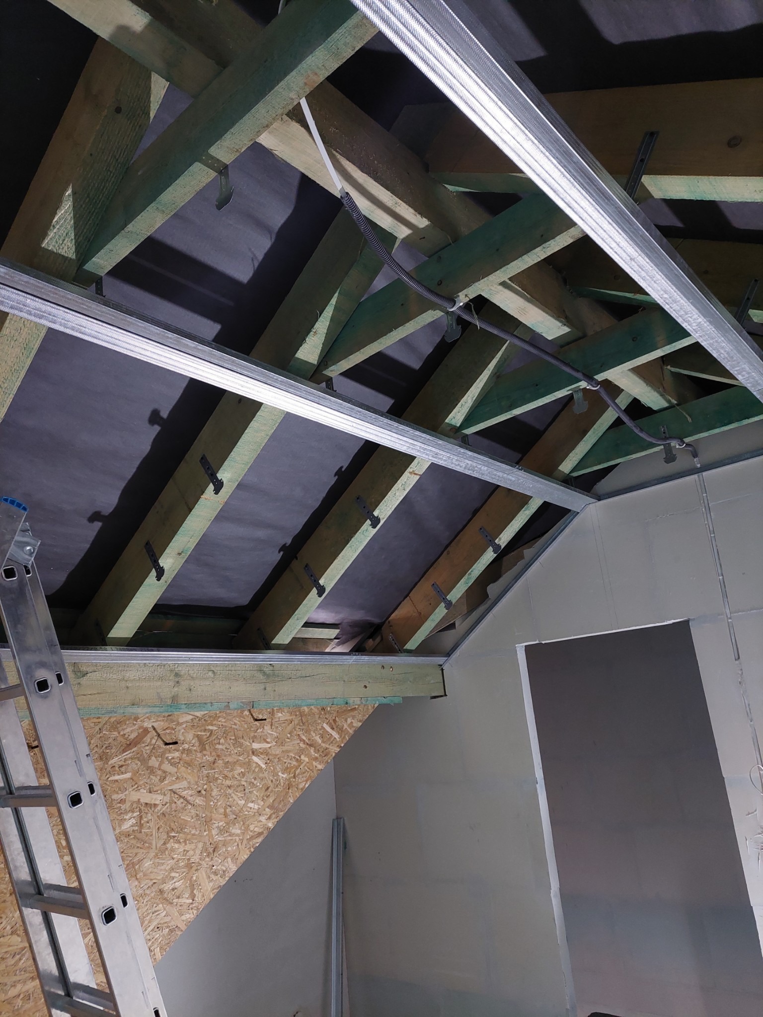 Montaż metalowego stelaża pod płytę karton gips na drewnianej konstrukcji dachu skośnego, widoczna drabina aluminiowa i fragment ściany z płyt OSB.