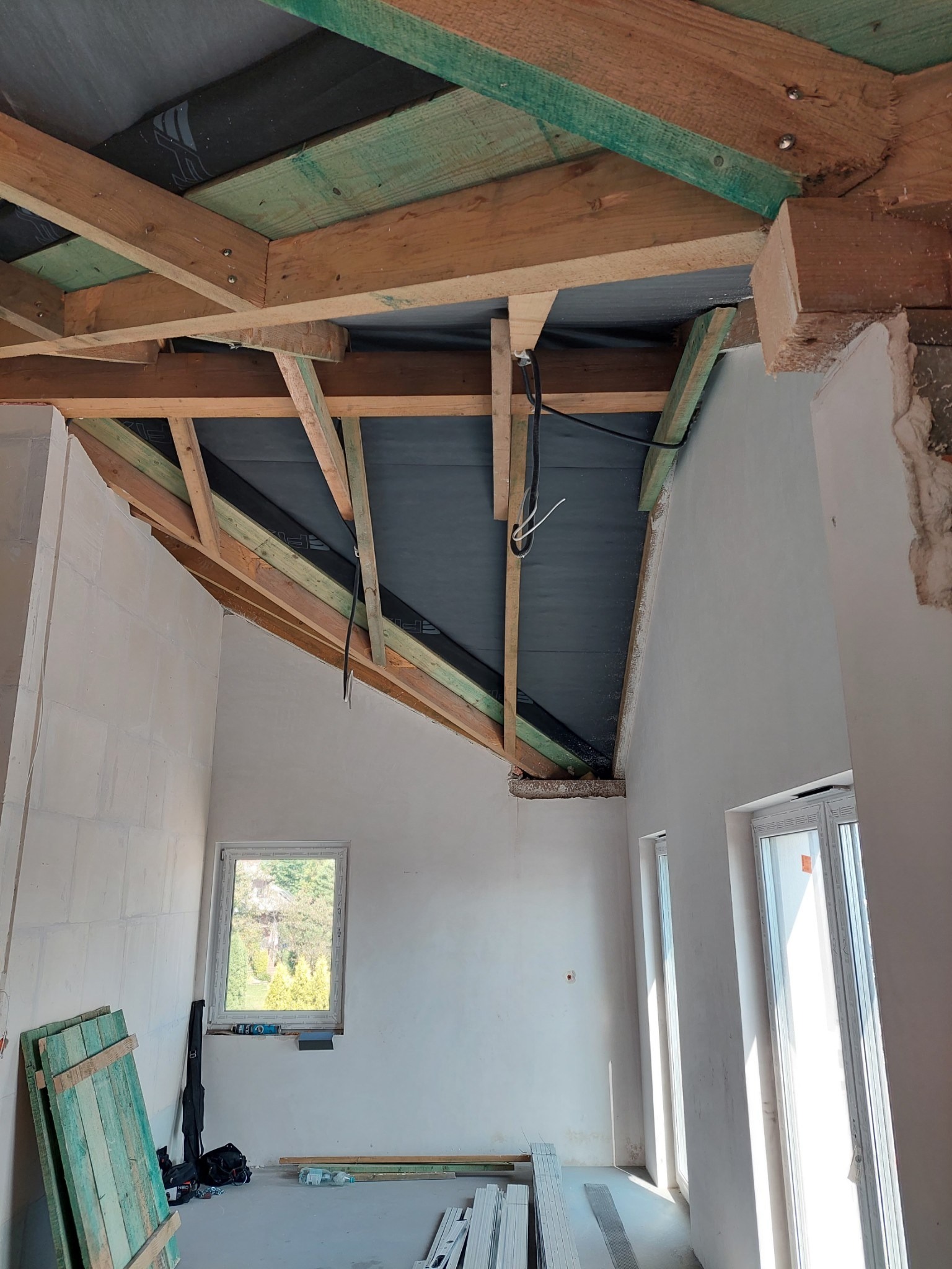 Poddasze w trakcie remontu: widoczne drewniane elementy konstrukcyjne dachu z czarną folią, ściany przygotowane do wykończenia, okna, oraz materiały budowlane na podłodze.