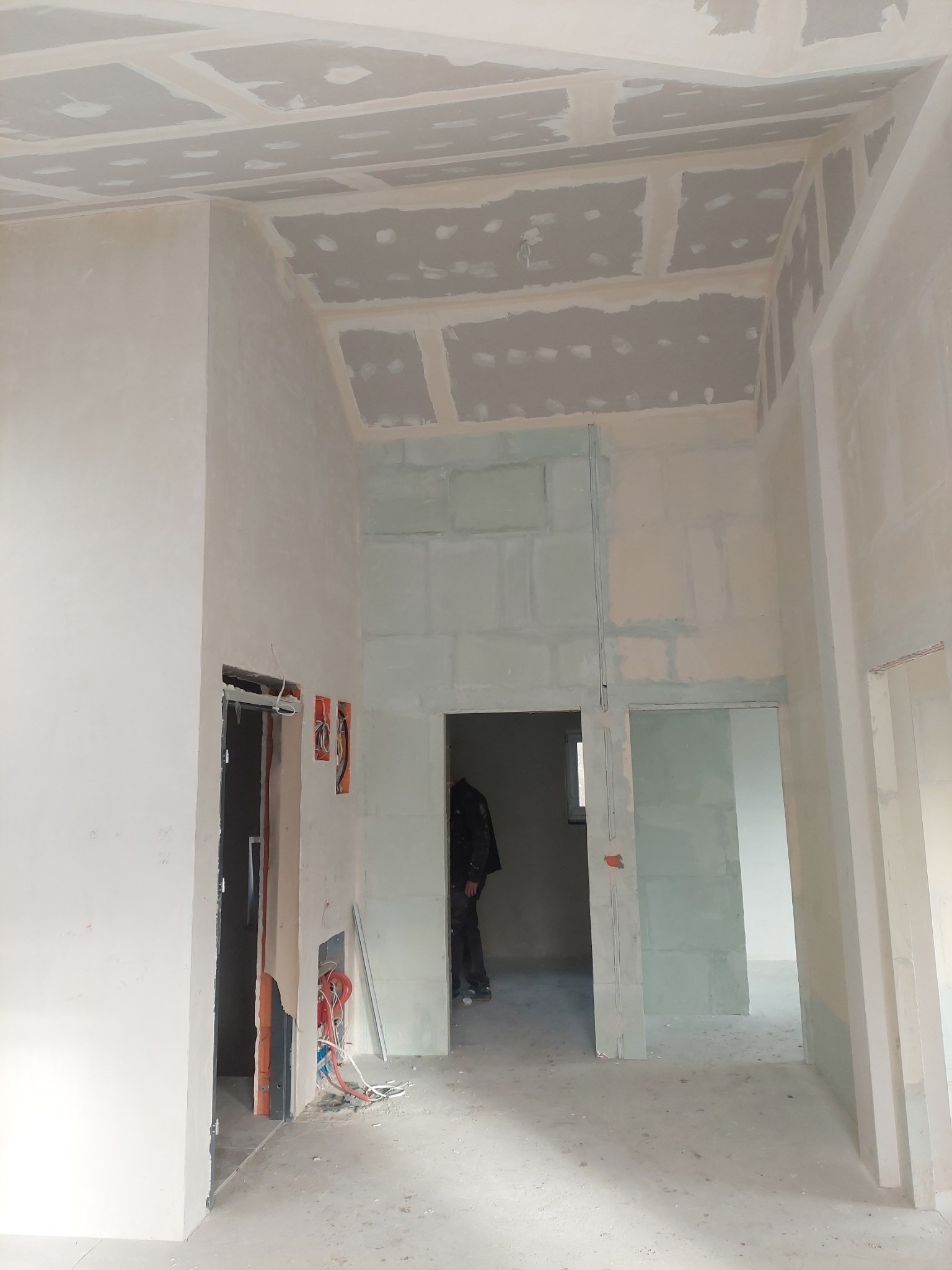 Wnętrze w trakcie prac wykończeniowych: widoczne płyty kartonowo-gipsowe na suficie i ścianach, niezabezpieczone przewody elektryczne wystające ze ściany, fragment sylwetki osoby w tle.