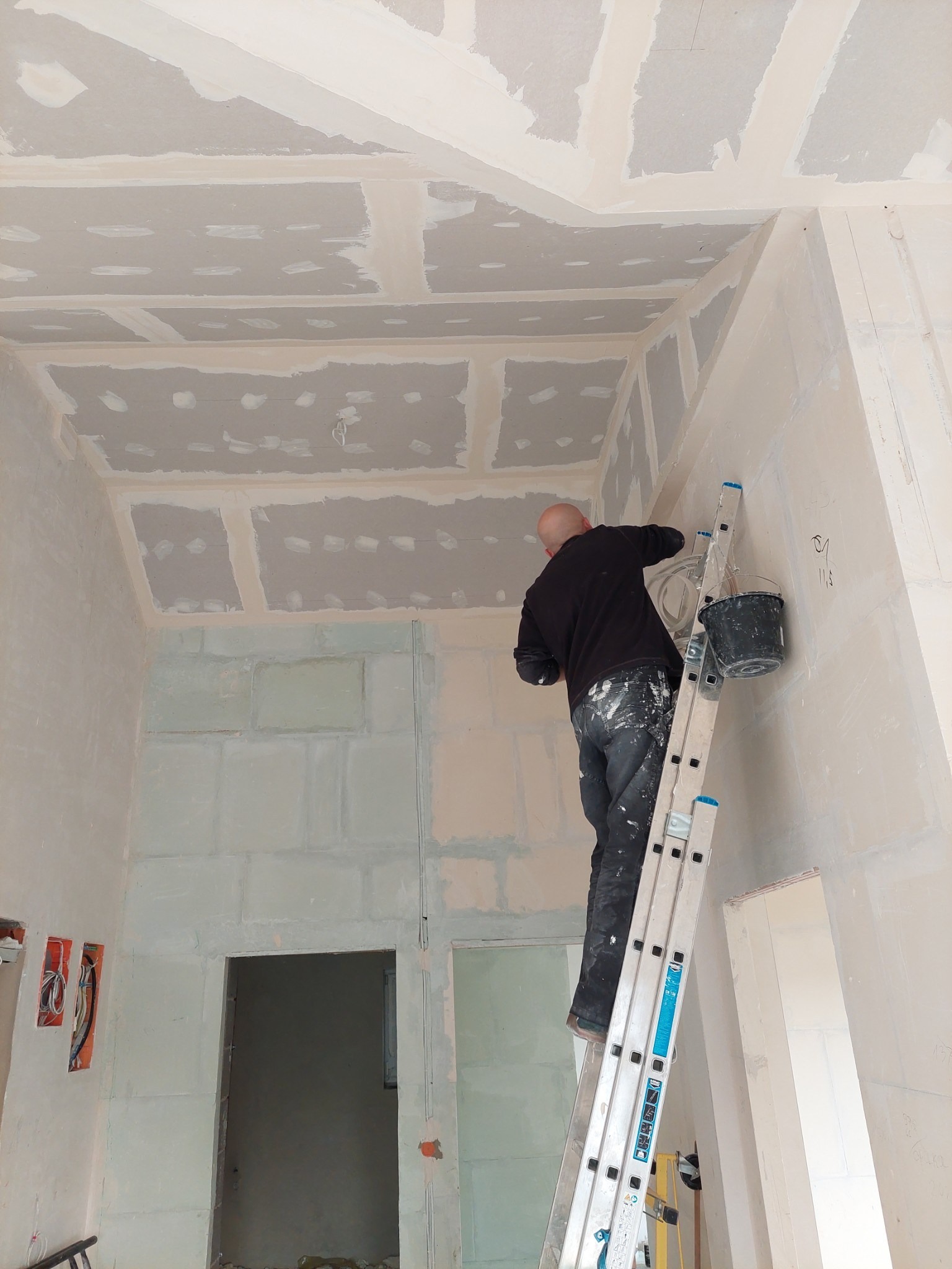 Pracownik na drabinie szpachluje narożnik ściany z płyt kartonowo-gipsowych, widoczny sufit z płytami i wiadro z masą szpachlową.