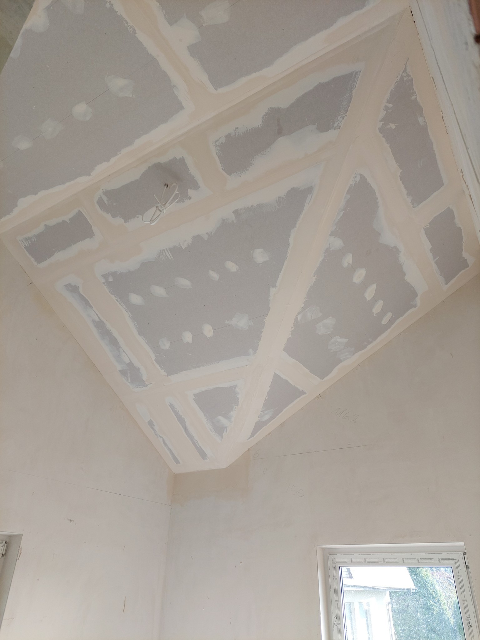 Sufit z płyt kartonowo-gipsowych, częściowo zaszpachlowany, widoczne łączenia płyt i instalacja elektryczna, ściany przygotowane do dalszych prac wykończeniowych.