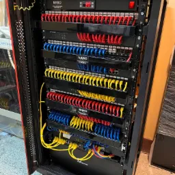 Szafa rack z zamontowanym osprzętem sieciowym: patch panele z kolorowymi kablami Ethernet (czerwone, niebieskie, żółte), switche TP-Link i DrayTek, listwa zasilająca z wyświetlaczem temperatury...