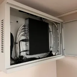 Zamknięta szafka natynkowa z urządzeniem rejestrującym i uporządkowanymi kablami sieciowymi, HDMI, USB, zainstalowana przy suficie na jasnej ścianie.