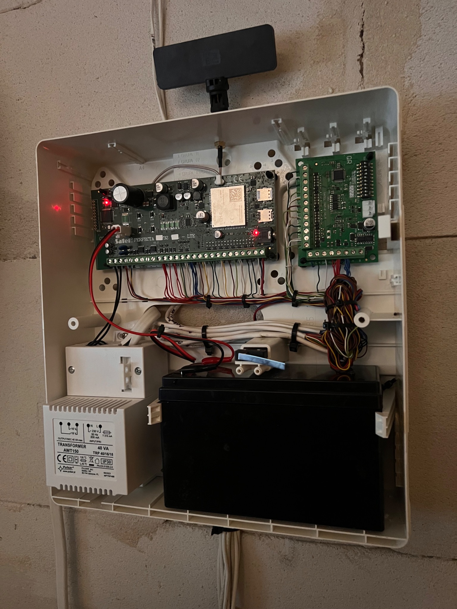 Otwarta obudowa centrali alarmowej z widocznymi modułami elektronicznymi, transformatorem Pulsar AWT150, akumulatorem i uporządkowanymi wiązkami przewodów na tle surowej ściany.