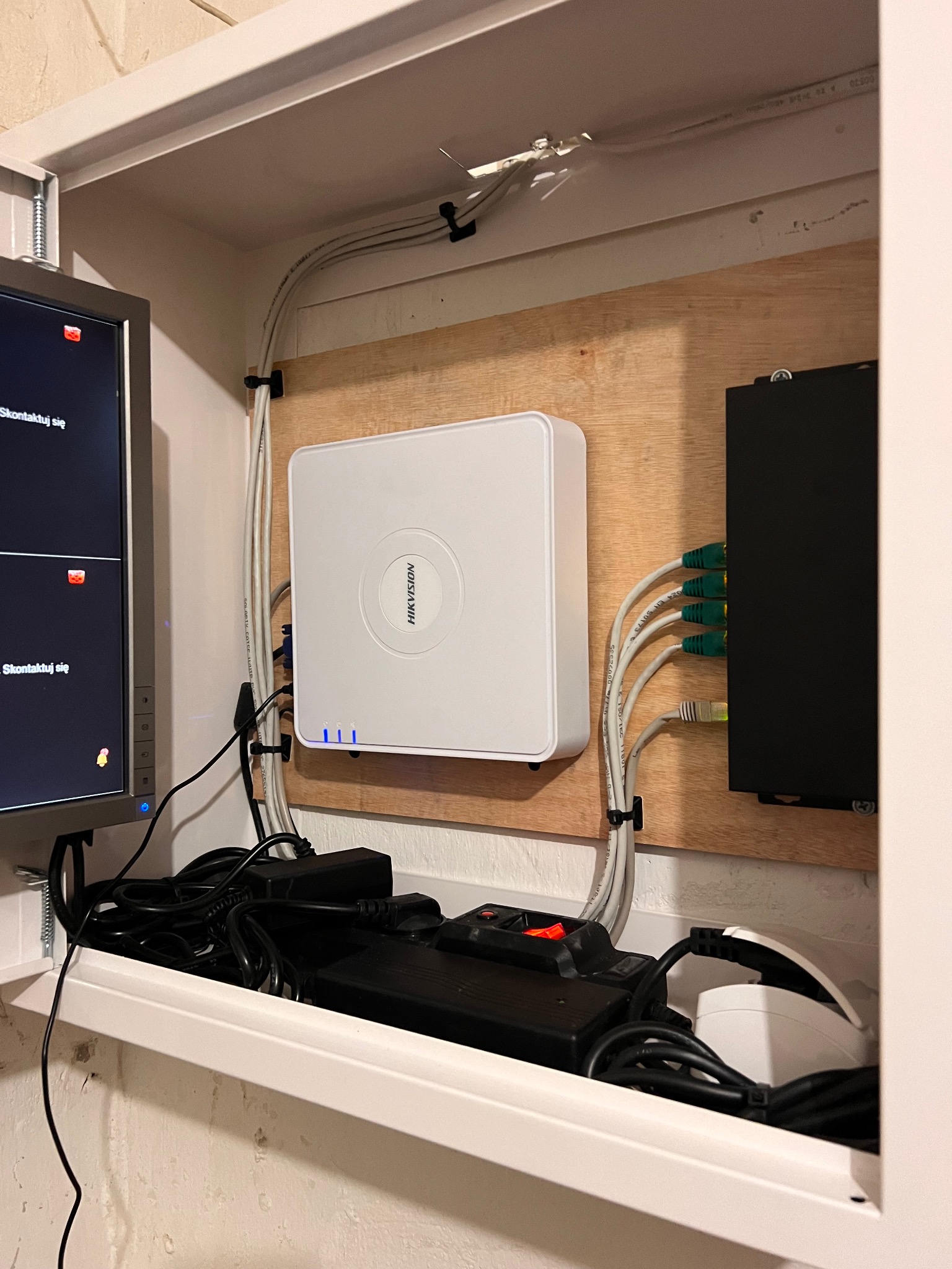 Zamknięta szafka z zamontowanym rejestratorem Hikvision z trzema świecącymi diodami, czarną skrzynką rozdzielczą z podłączonymi kablami Ethernet, monitorem z widocznymi ikonami powiadomień,...
