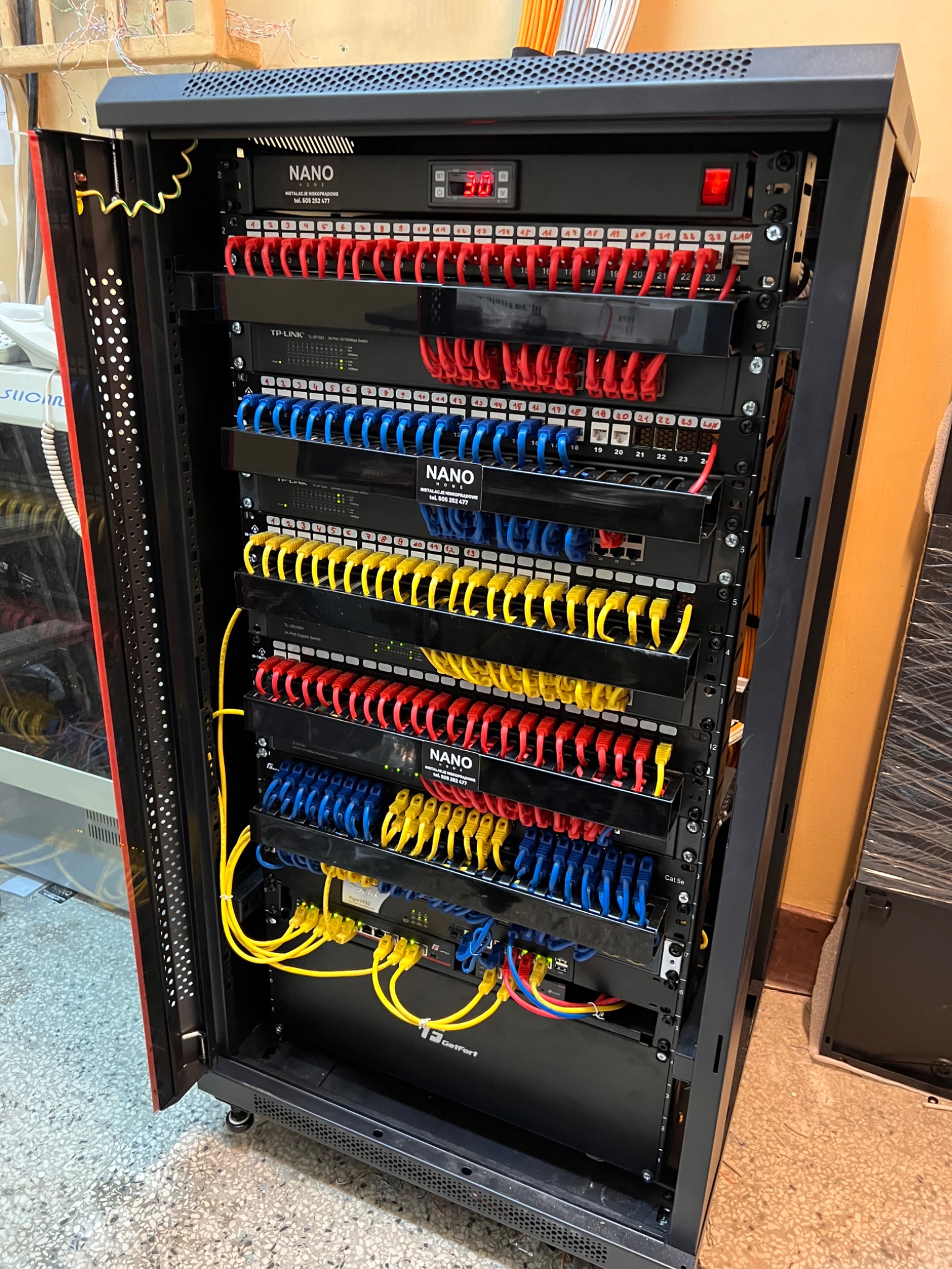 Szafa rack z zamontowanym osprzętem sieciowym: patch panele z kolorowymi kablami Ethernet (czerwone, niebieskie, żółte), switche TP-Link i DrayTek, listwa zasilająca z wyświetlaczem temperatury...