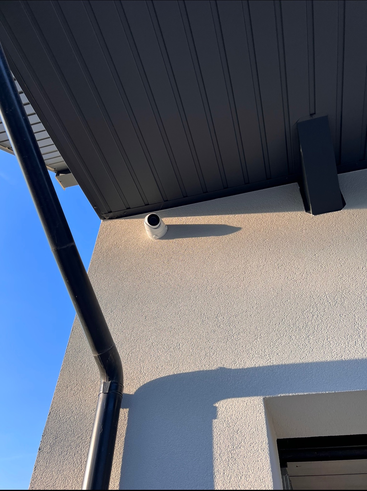 Biała kamera monitoringu zamontowana na elewacji domu pod czarnym dachem i rynną, widok z dołu w słoneczny dzień.