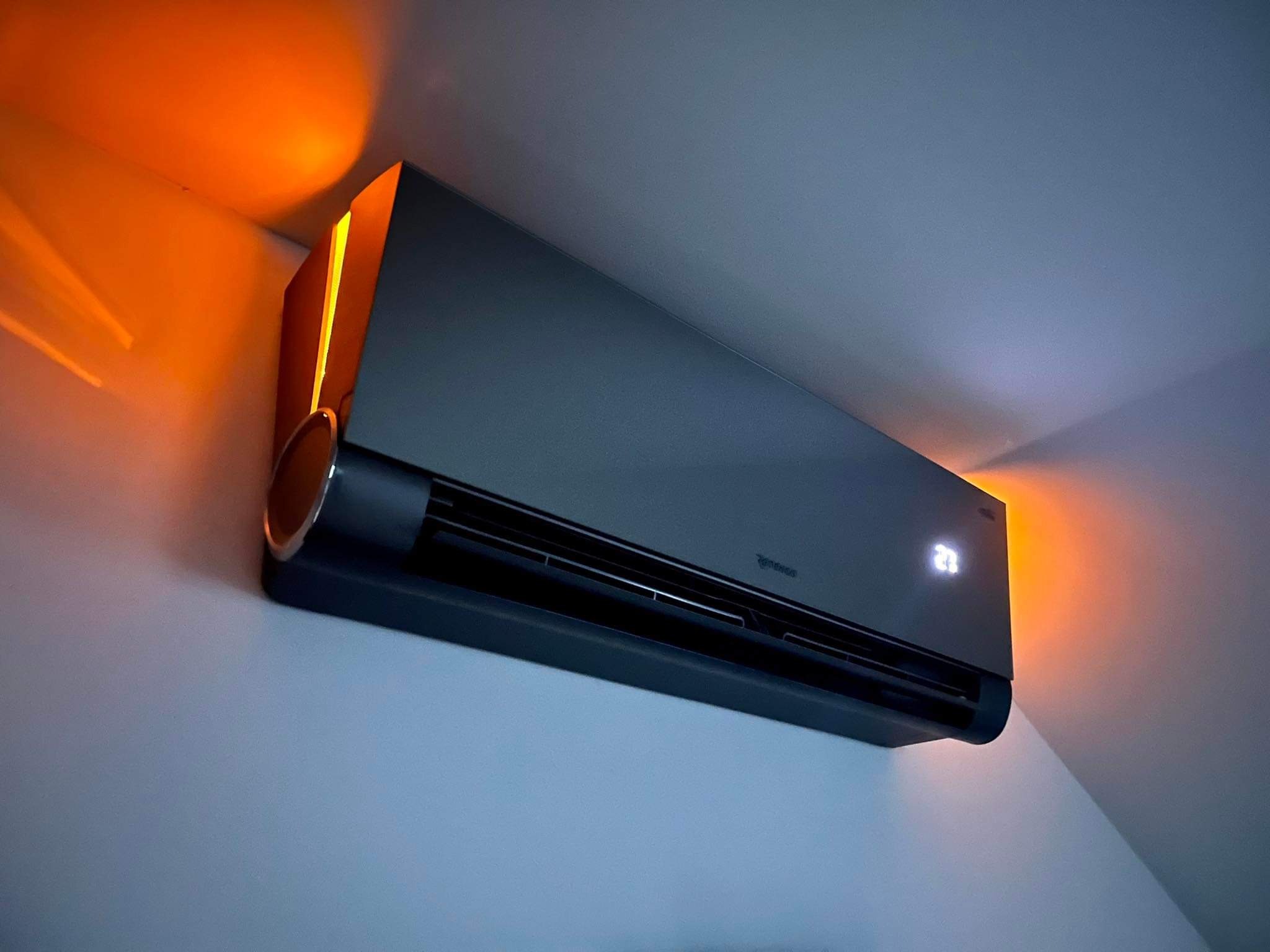 Czarny klimatyzator ścienny z podświetleniem LED w kolorze pomarańczowym, wyświetlający temperaturę 23 stopnie Celsjusza, zamontowany na białej ścianie.