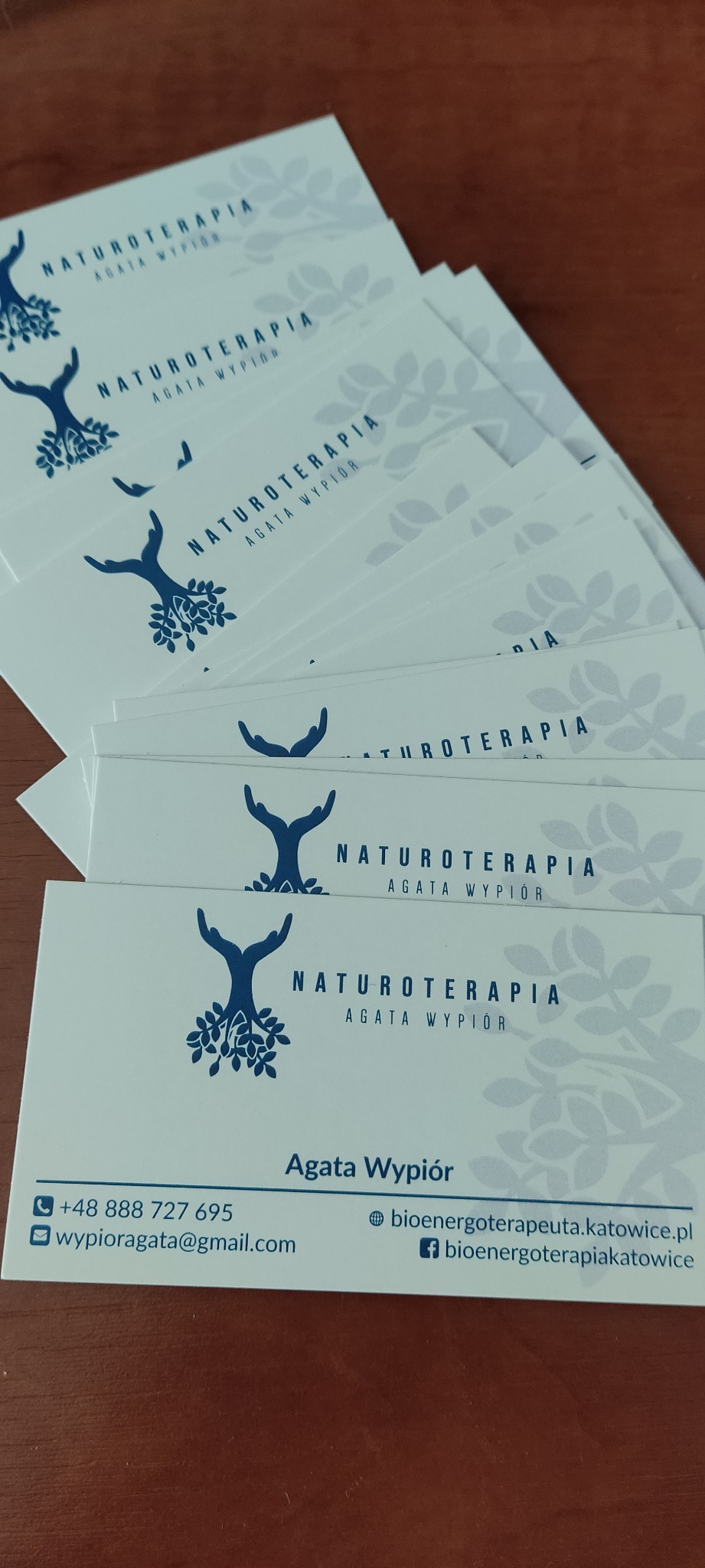 Stos wizytówek z logo firmy Naturoterapia Agata Wypiór, na wizytówkach grafika drzewa wyrastającego z dłoni, dane kontaktowe.