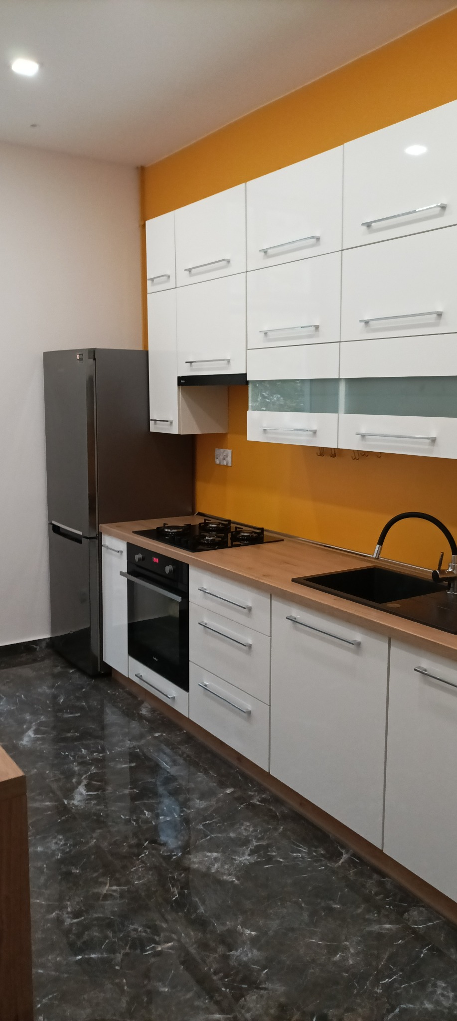 Nowoczesna kuchnia z białymi szafkami, drewnianym blatem i marmurową podłogą, akcentowana pomarańczową ścianą.