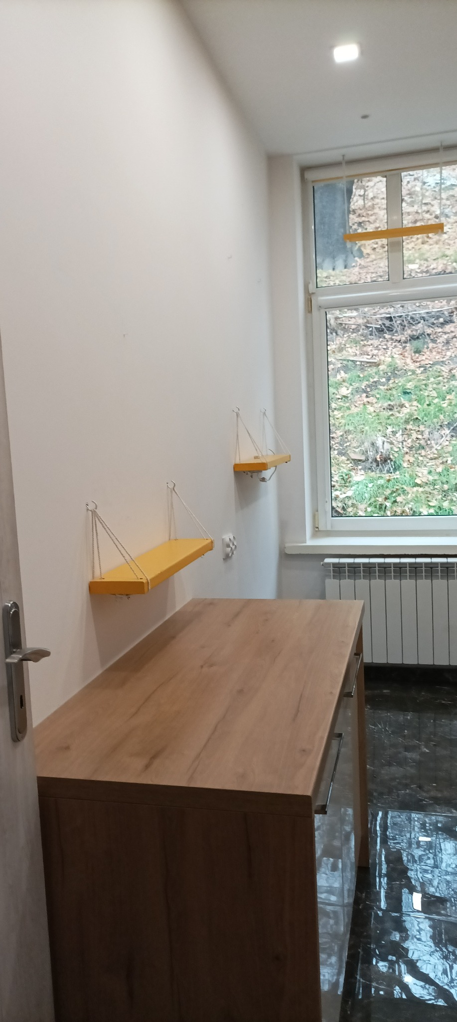 Jasne wnętrze z drewnianym blatem, dwiema żółtymi półkami wiszącymi na linkach i widokiem za oknem na jesienny ogród. Blat ma widoczne szuflady z metalowymi uchwytami.