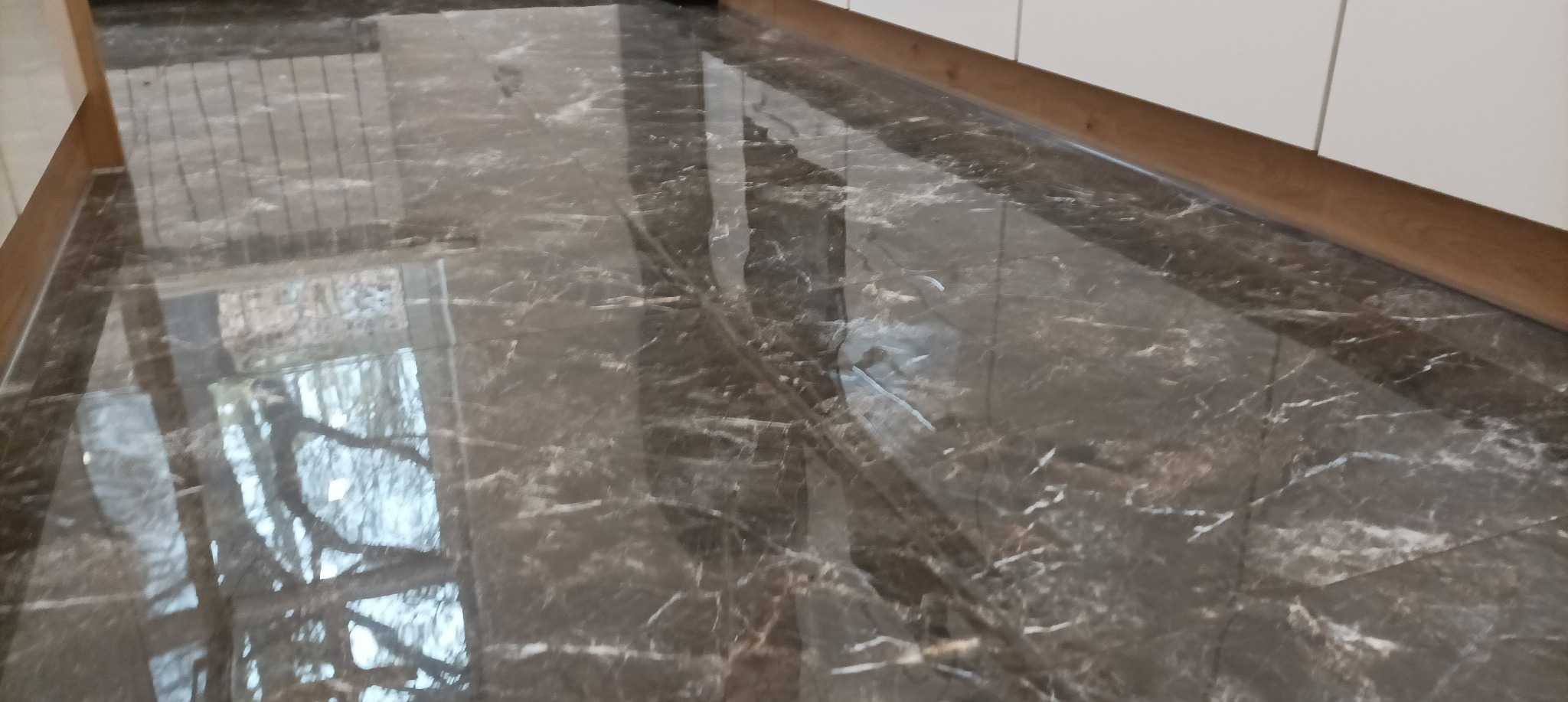 Polerowana, brązowa podłoga z płytek imitujących marmur, z widocznym odbiciem okna i drzew, ułożona przy listwie przypodłogowej.