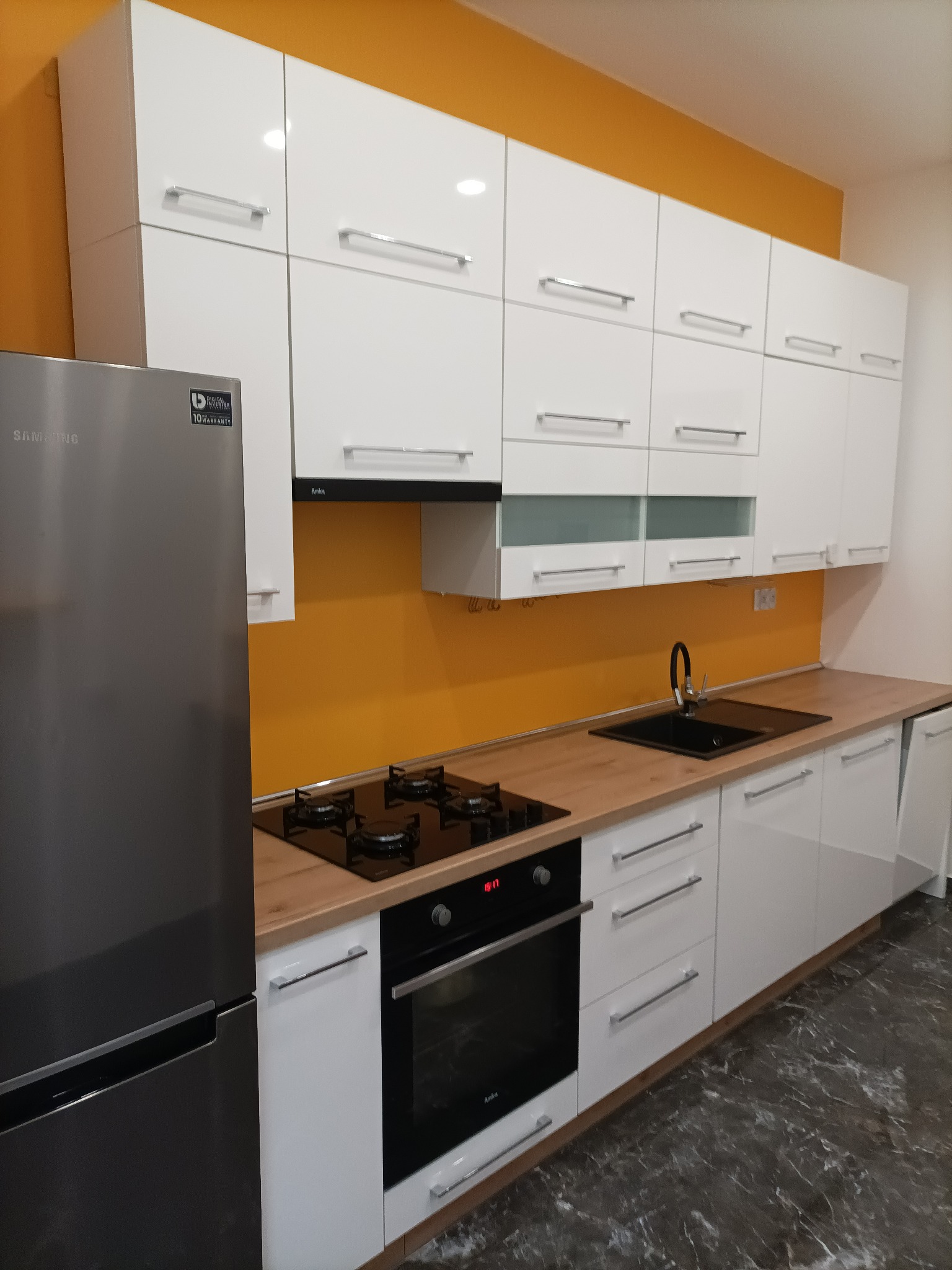 Nowoczesna kuchnia z białymi, połyskującymi szafkami, drewnianym blatem, czarnym zlewem i płytą grzewczą, z żółtym panelem na ścianie i lodówką Samsung po lewej stronie.