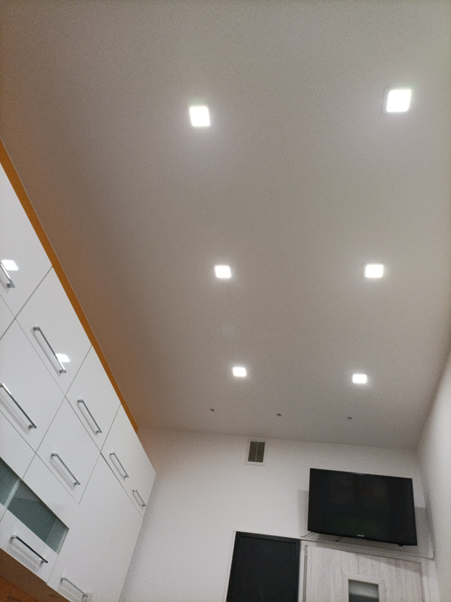 Biały sufit z równomiernie rozmieszczonymi kwadratowymi lampami LED, widok z dołu, fragment białych szafek z szufladami po lewej stronie, telewizor na ścianie w tle.