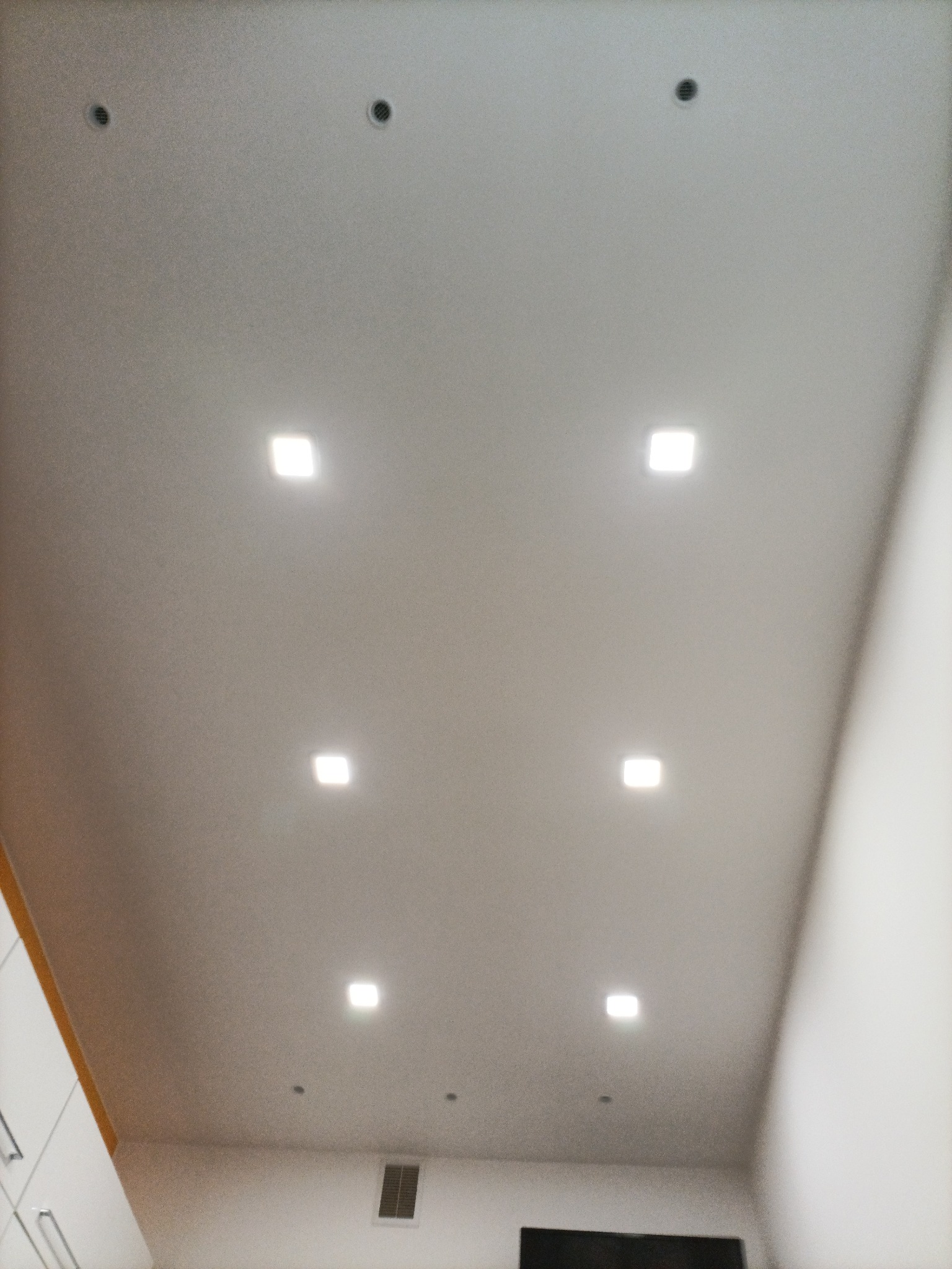 Biały sufit z równomiernie rozmieszczonymi kwadratowymi lampami LED i okrągłymi punktami wentylacyjnymi, widok z dołu.