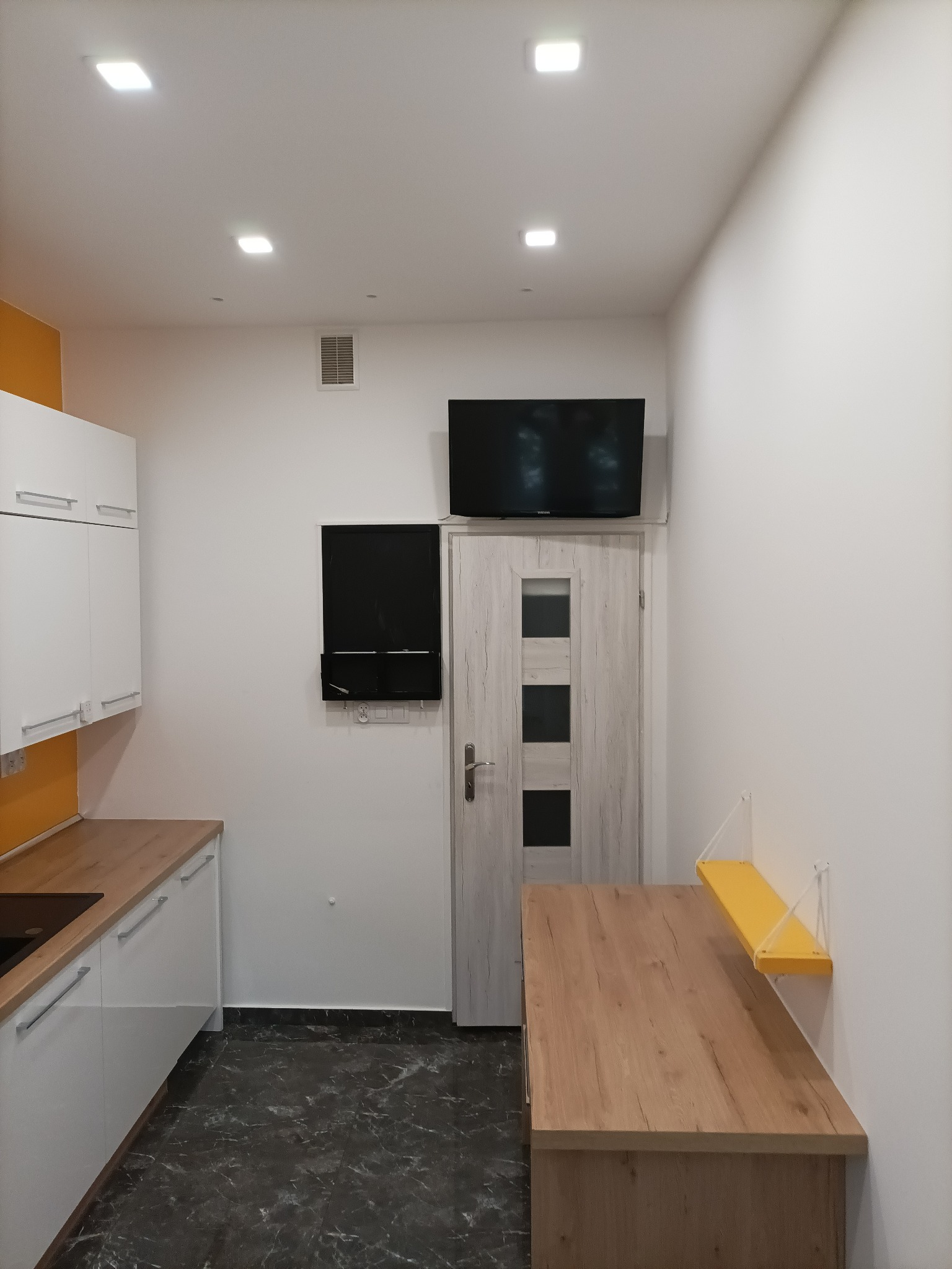 Nowoczesna kuchnia z białymi szafkami, drewnianym blatem i stołem, czarną płytą grzewczą, czarnym telewizorem nad drzwiami i marmurkową podłogą.