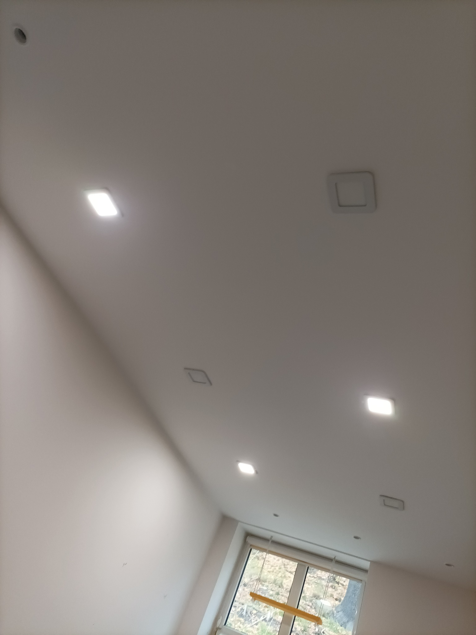 Biały sufit z zamontowanymi kwadratowymi lampami LED w Gubinie, widok z dołu, okno w tle.