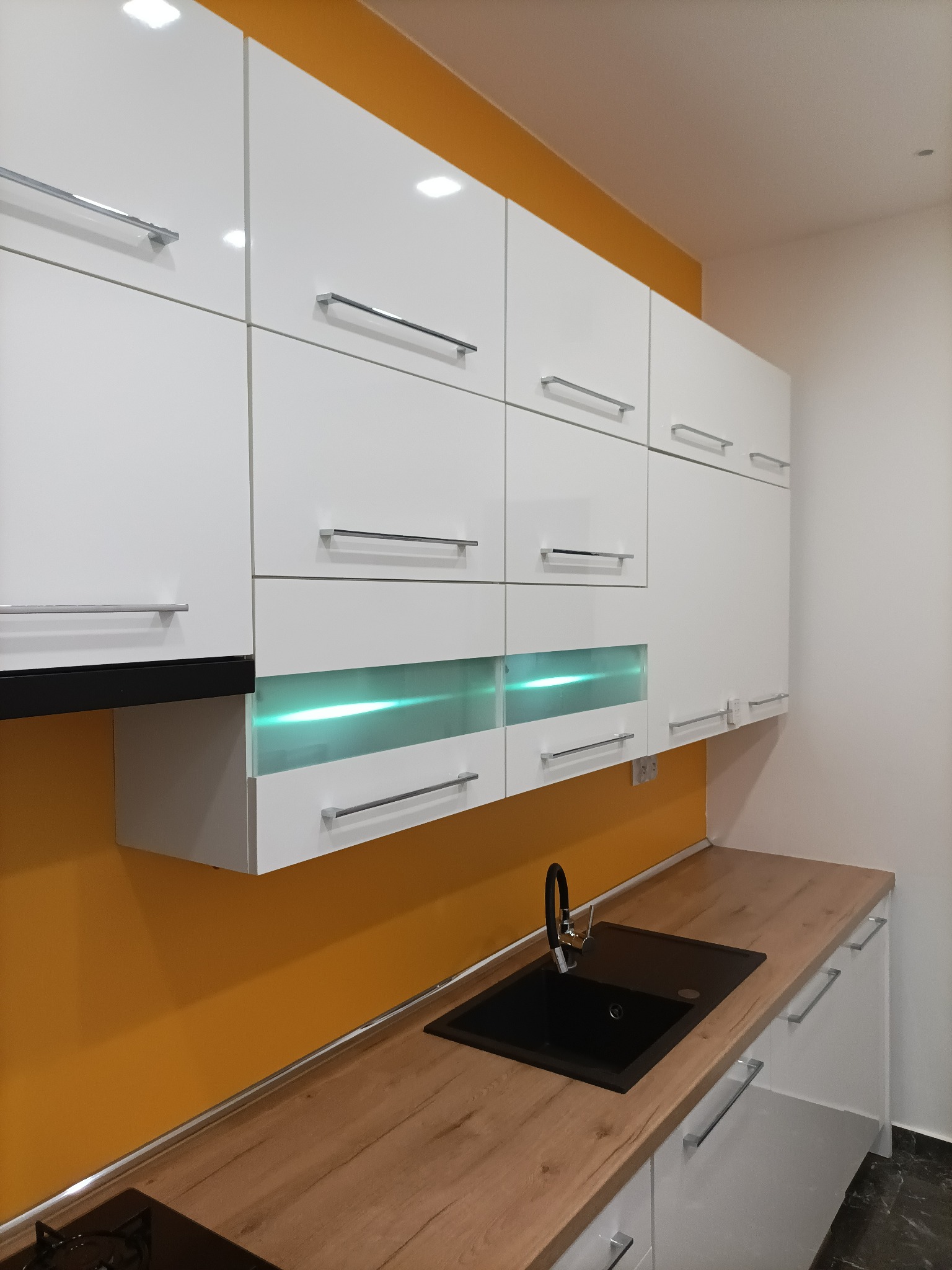 Nowoczesna kuchnia z białymi szafkami, drewnianym blatem i czarnym zlewozmywakiem, akcentowana pomarańczową ścianą i podświetlanymi szafkami.