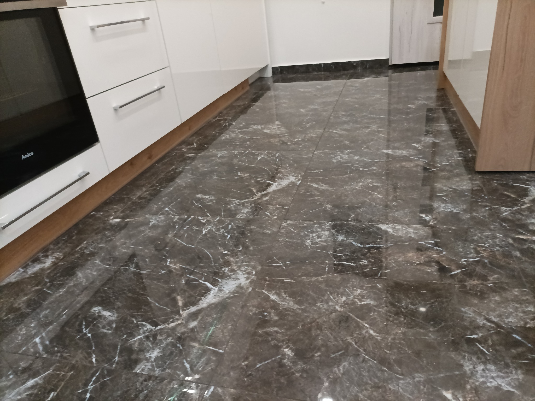 Błyszcząca, ciemna podłoga z marmurowym wzorem w nowocześnie wykończonej kuchni z białymi szafkami i stalowymi uchwytami.
