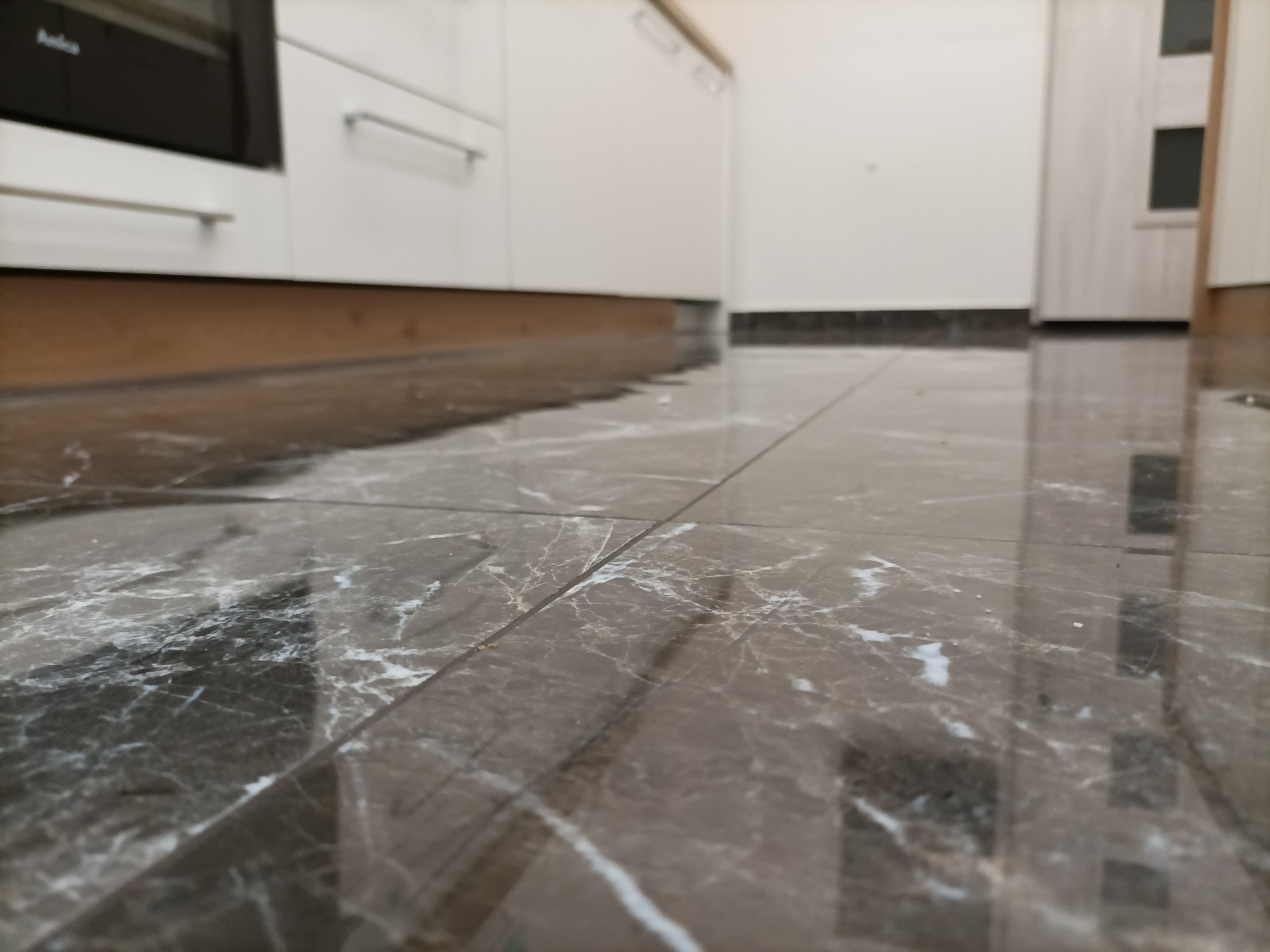 Ułożona podłoga z płytek imitujących marmur w kuchni z białymi szafkami, widoczne fugi i odbicia światła.