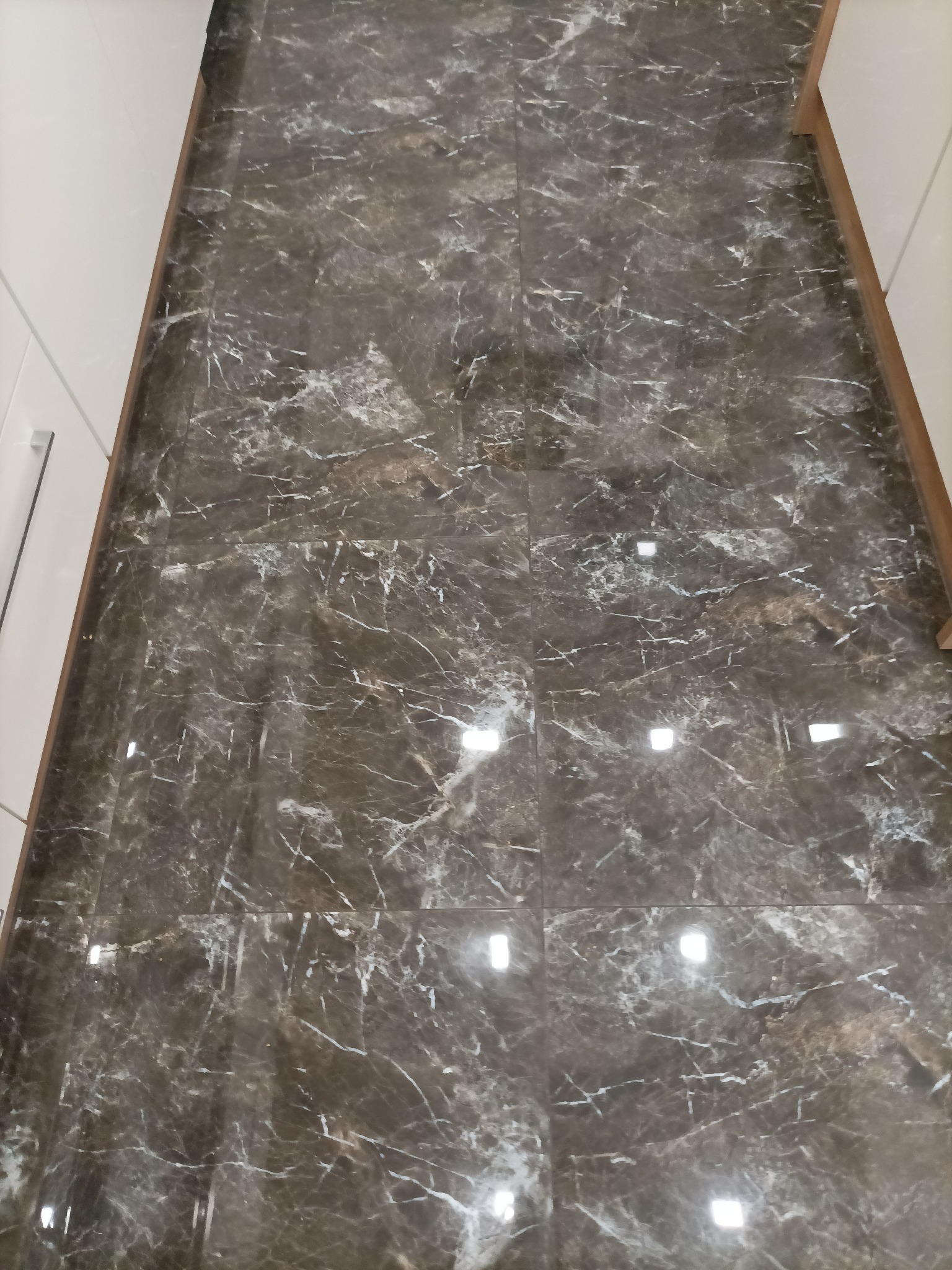 Ułożona, ciemna, marmurkowa podłoga z płytek, z widocznymi odbiciami światła, przylegająca do białych i drewnianych elementów wykończeniowych.