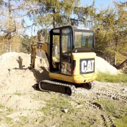 Żółta minikoparka marki CAT na placu budowy, z widocznymi śladami gąsienic na ziemi i stertą piasku w tle, z lasem iglastym i błękitnym niebem w tle.