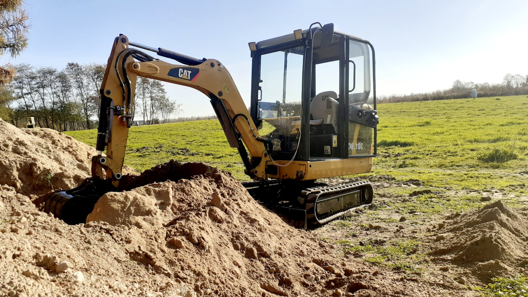 Żółta minikoparka CAT 301.8C w trakcie prac ziemnych na zielonym polu, widoczny wykop i usypana pryzma ziemi, błękitne niebo w tle.