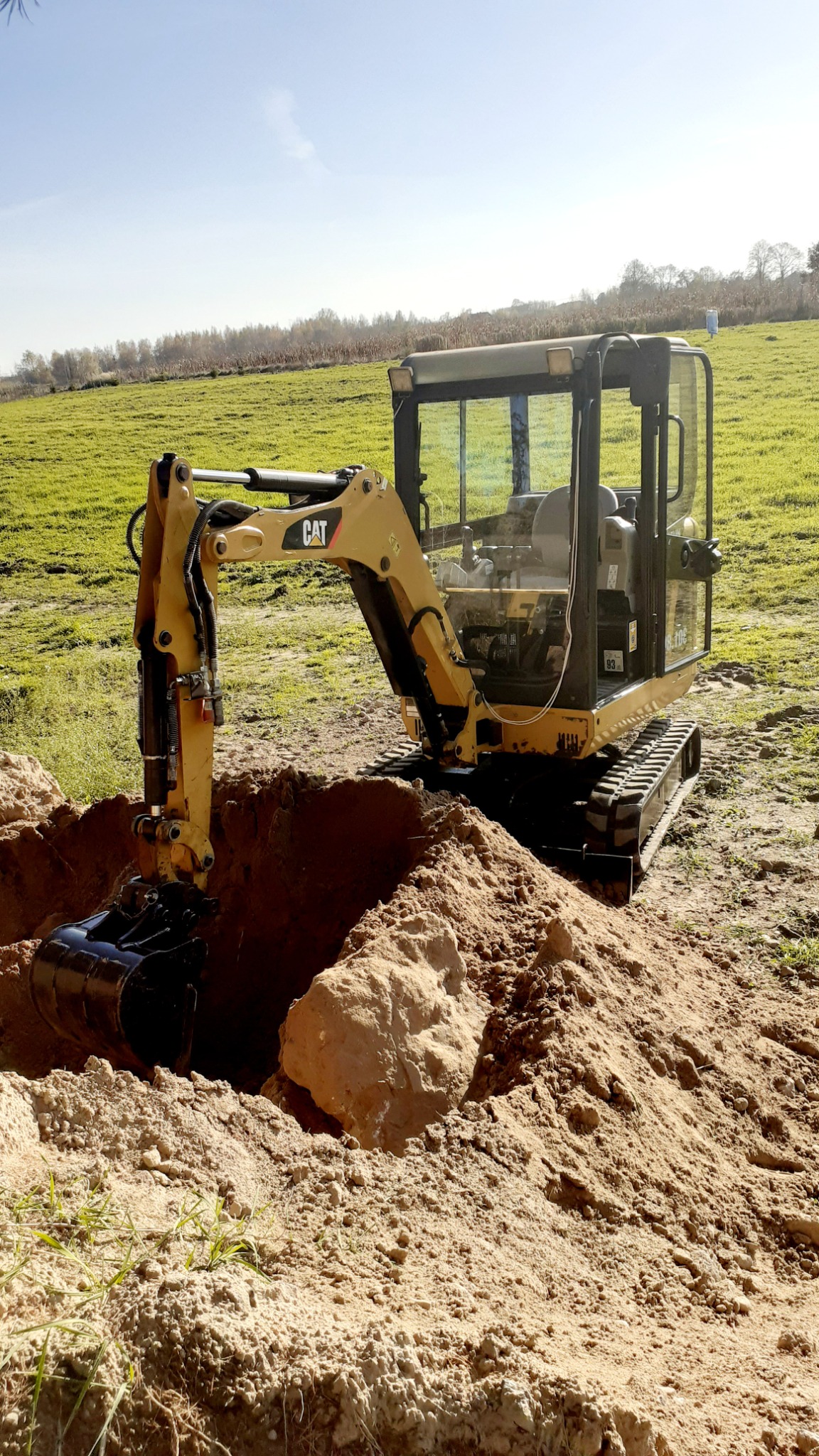 Żółta minikoparka CAT podczas prac ziemnych, widoczny wykop w jasnej ziemi z dużym kamieniem, zielone pole w tle.