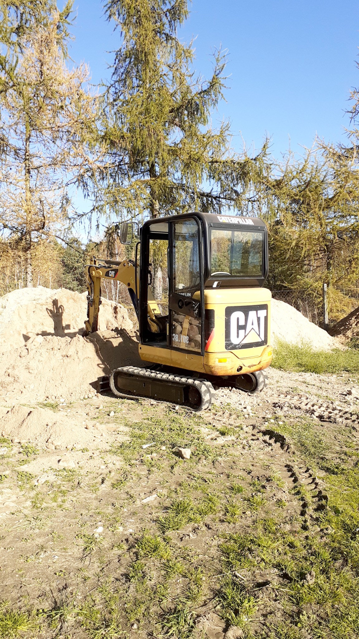 Żółta minikoparka marki CAT na placu budowy, z widocznymi śladami gąsienic na ziemi i stertą piasku w tle, z lasem iglastym i błękitnym niebem w tle.