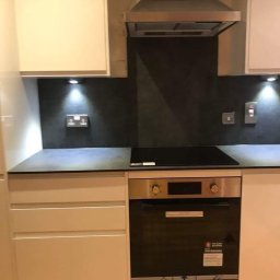 AspireDesign - Fragment wykończonej kuchni z białymi szafkami, czarnym blatem, indukcyjną płytą grzewczą i piekarnikiem, oświetleniem podszafkowym i metalowym okapem nad czarną ścianą między szafkami.