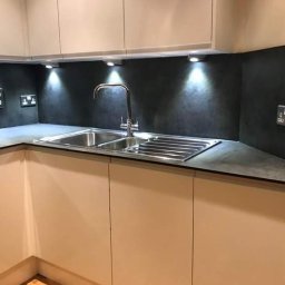 AspireDesign - Nowoczesna kuchnia z podświetlanym blatem i zlewozmywakiem dwukomorowym w odcieniach szarości i beżu, widoczne gniazdka elektryczne na ścianie.