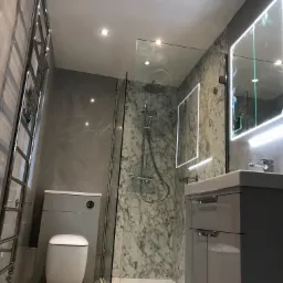 Nowoczesna łazienka w szarościach z marmurową ścianą prysznicową, podświetlanym lustrem, grzejnikiem drabinkowym i wiszącą toaletą.