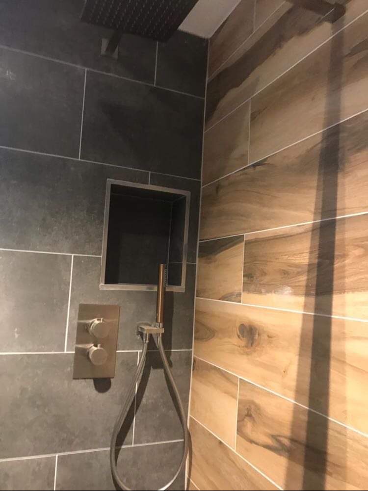 Nowoczesna kabina prysznicowa z szarymi płytkami imitującymi kamień, drewnopodobnymi panelami, wnęką na kosmetyki i metalową armaturą w odcieniach brązu.