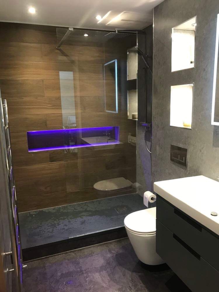 Nowoczesna łazienka z kabiną prysznicową typu walk-in, podświetlaną wnęką z fioletowym światłem, szarą umywalką z szafką i wiszącą toaletą.