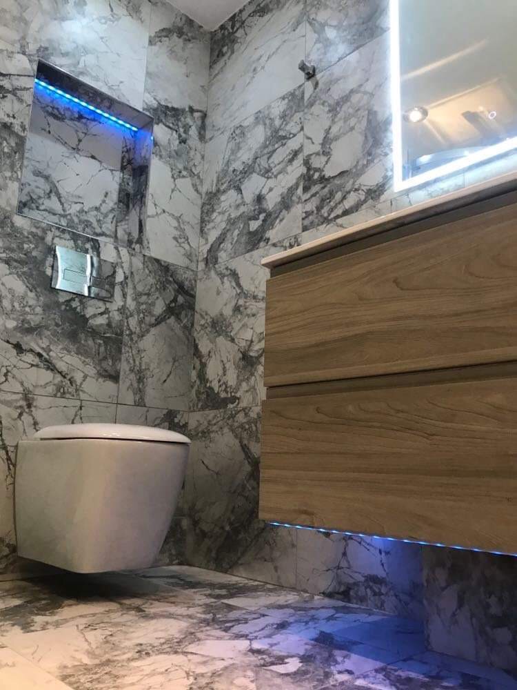 Nowoczesna łazienka z marmurowymi płytkami, podwieszaną toaletą i szafką pod umywalką z podświetleniem LED, tworząca elegancki i minimalistyczny design.