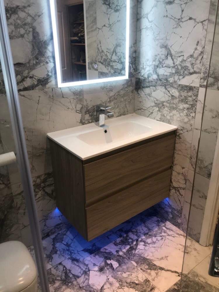 Nowoczesna łazienka z umywalką nablatową na szafce z drewnopodobnym fornirem, podświetleniem LED od dołu i lustrem z oświetleniem LED, marmurowe płytki na ścianach i podłodze.