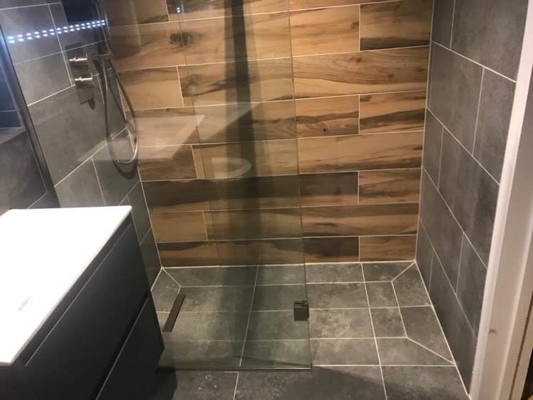 Nowoczesna kabina prysznicowa z drewnopodobnymi płytkami na ścianie, szare płytki podłogowe i szklane drzwi z minimalistycznym zawiasem.