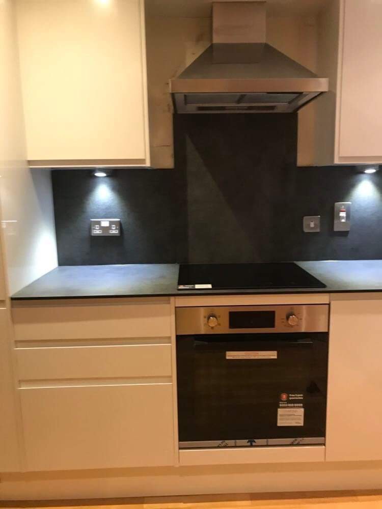 Fragment wykończonej kuchni z białymi szafkami, czarnym blatem, indukcyjną płytą grzewczą i piekarnikiem, oświetleniem podszafkowym i metalowym okapem nad czarną ścianą między szafkami.