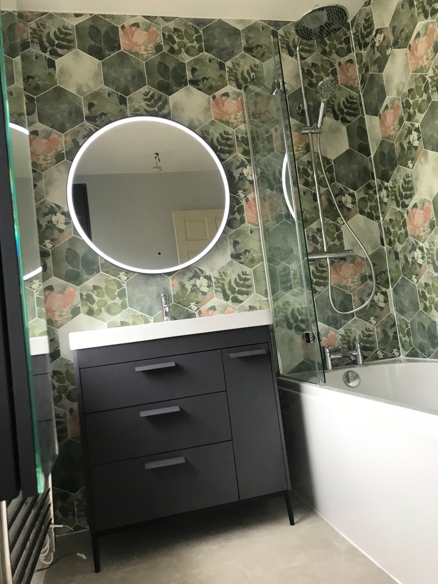 Wykończona łazienka z heksagonalnymi płytkami w roślinny wzór, okrągłe lustro z podświetleniem LED, czarna szafka pod umywalką i kabina prysznicowa nad wanną.