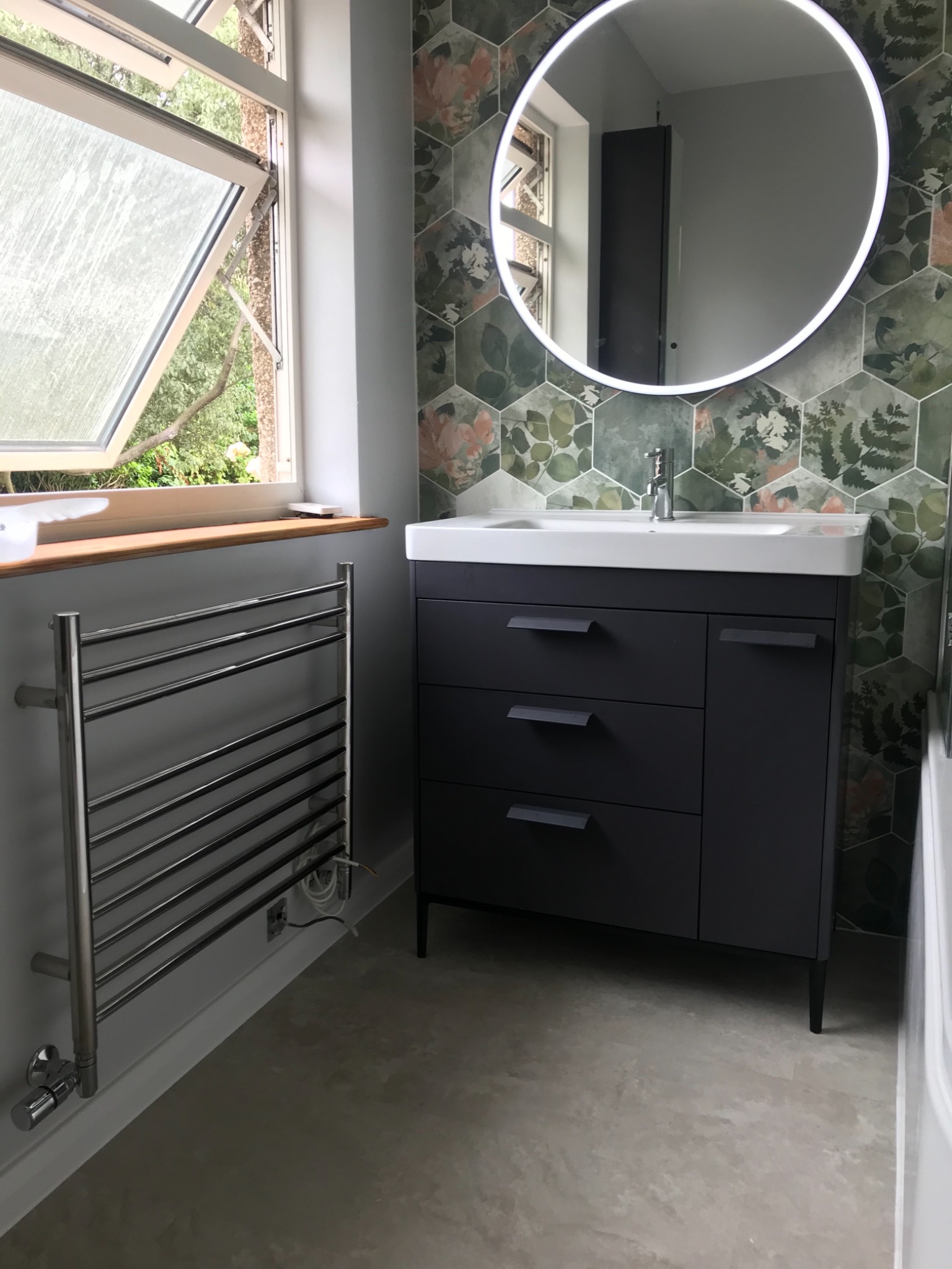 Nowoczesna łazienka z szarą szafką pod umywalką, okrągłym lustrem z podświetleniem LED, grzejnikiem drabinkowym i heksagonalnymi płytkami z motywem roślinnym.