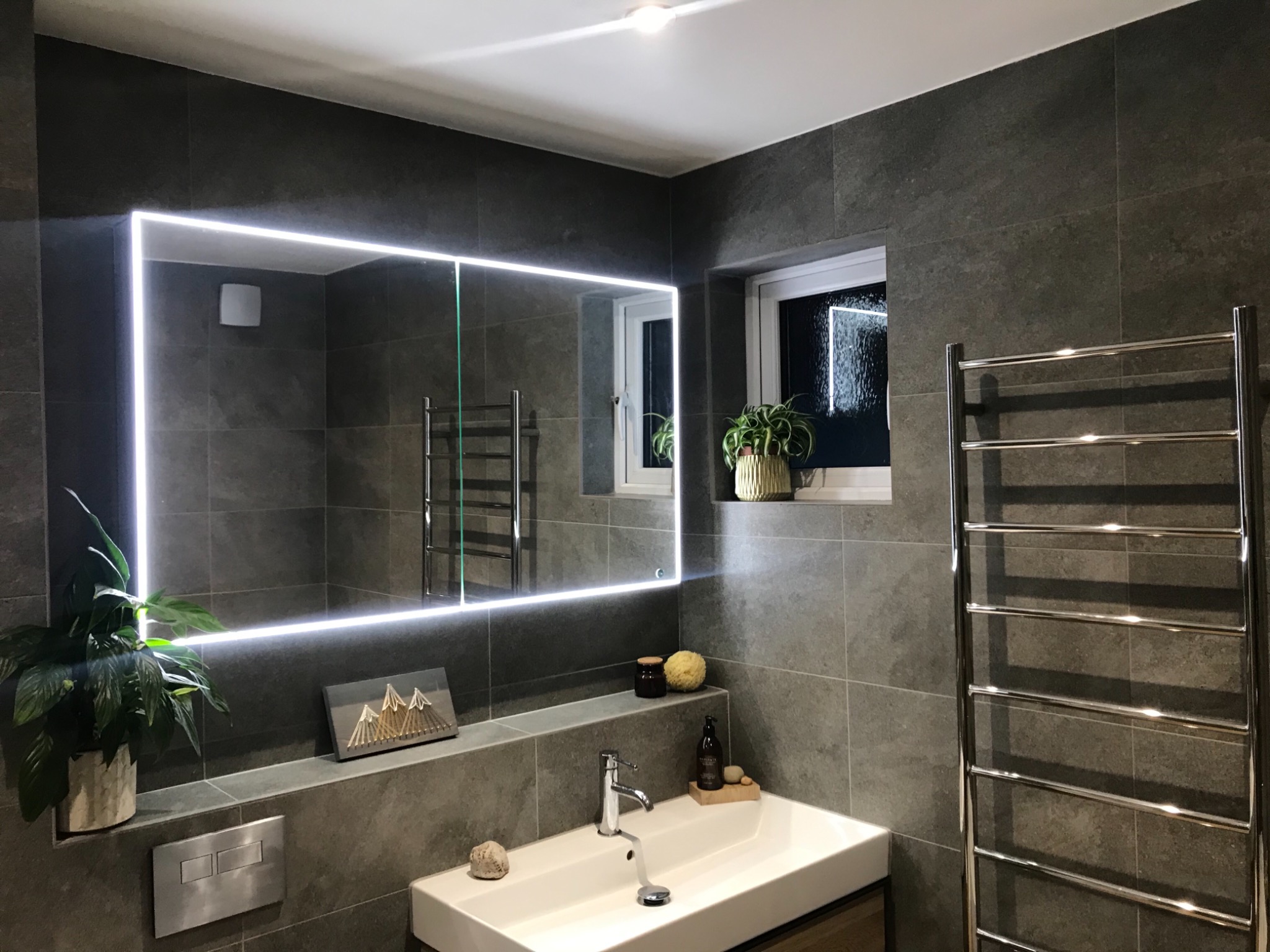 Nowoczesna łazienka z szarymi płytkami, podświetlanym lustrem, białą umywalką i grzejnikiem drabinkowym, uzupełniona roślinami i dekoracjami.