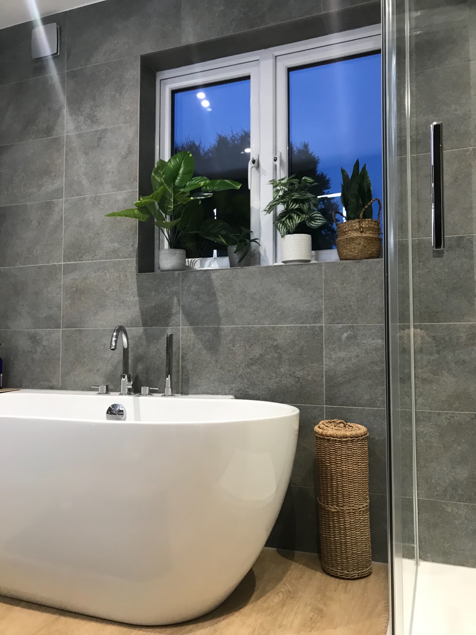 Nowoczesna łazienka z wolnostojącą wanną, szarą glazurą, roślinami doniczkowymi na parapecie okna i wiklinowym pojemnikiem na podłodze obok kabiny prysznicowej.