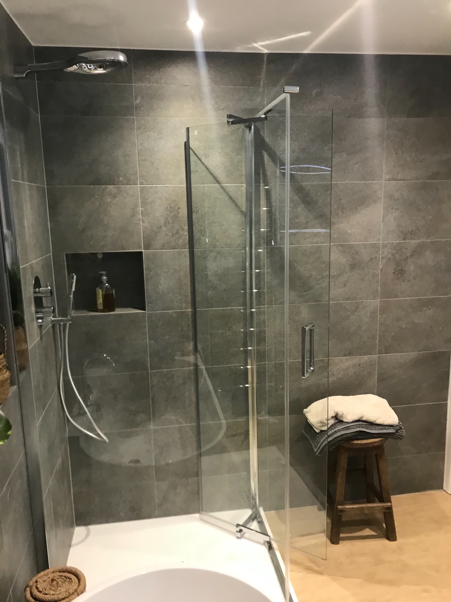 Nowoczesna kabina prysznicowa z szarymi płytkami, półką w ścianie z butelką mydła, deszczownicą i drewnianym stołkiem z ręcznikami w łazience.