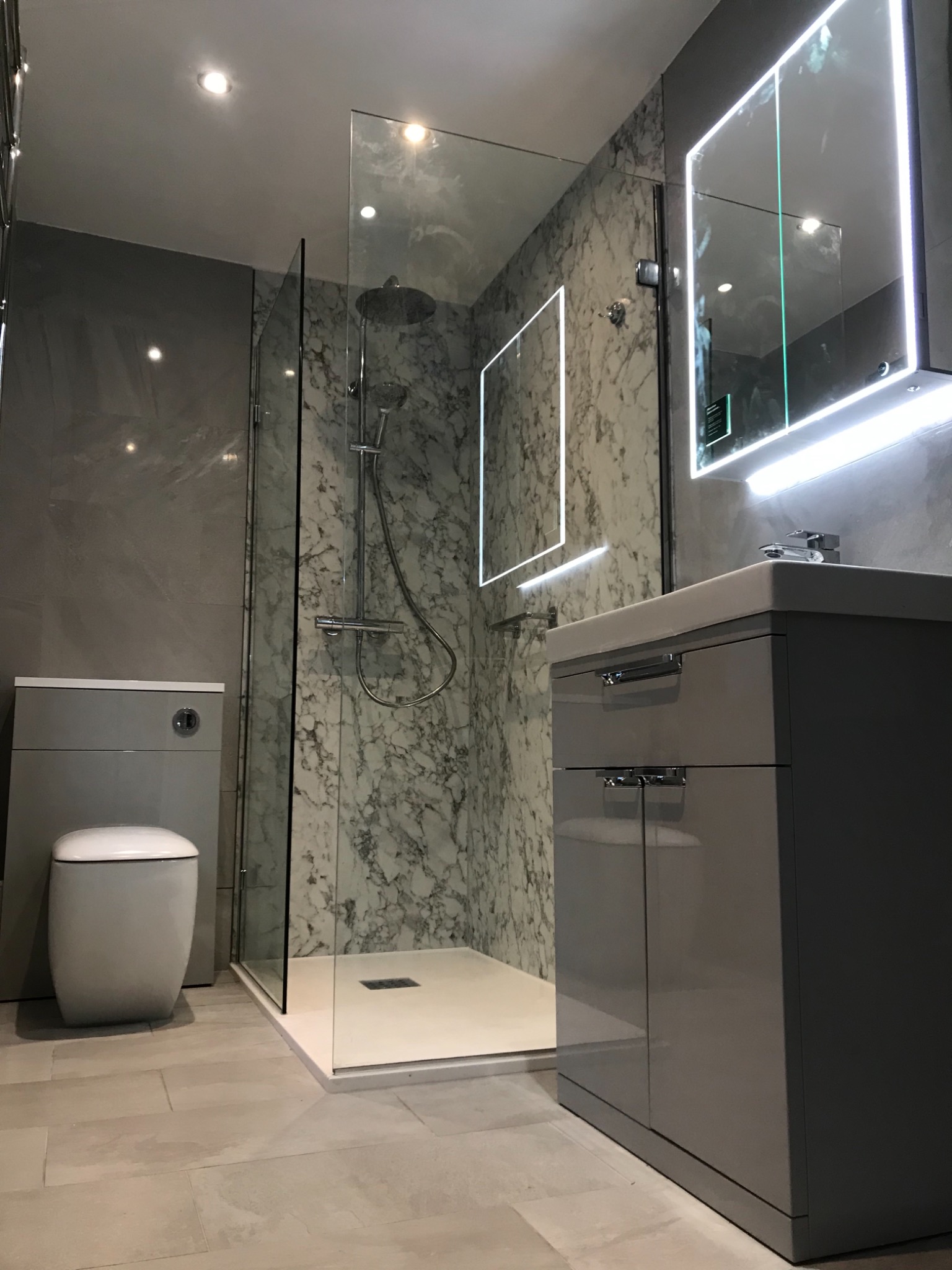 Nowoczesna łazienka z kabiną prysznicową typu walk-in, umywalką z szafką w kolorze szarym, toaletą w zabudowie i lustrami z podświetleniem LED.