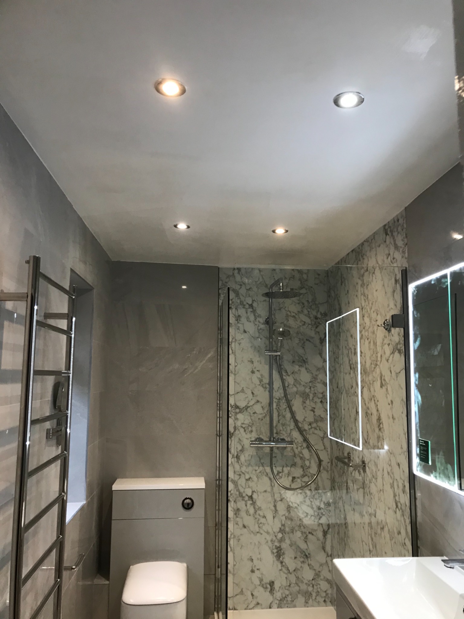 Nowoczesna łazienka z marmurowymi płytkami, szklaną kabiną prysznicową, podświetlanym lustrem i grzejnikiem drabinkowym, oświetlona punktowymi lampami sufitowymi.