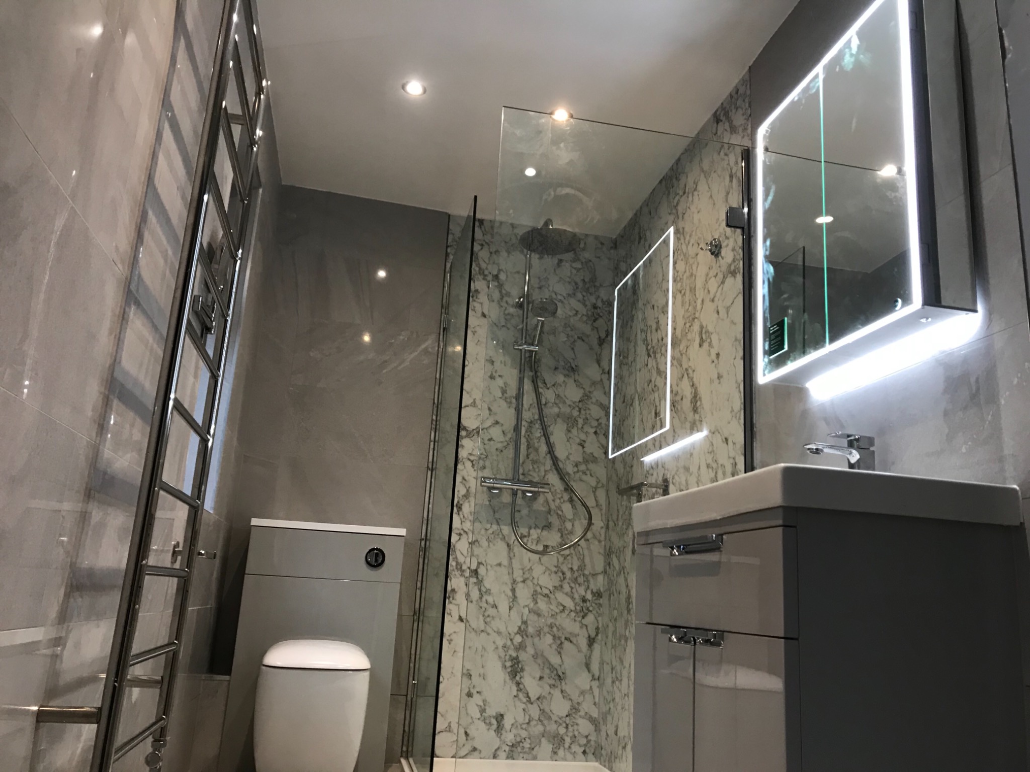 Nowoczesna łazienka w szarościach z marmurową ścianą prysznicową, podświetlanym lustrem, grzejnikiem drabinkowym i wiszącą toaletą.