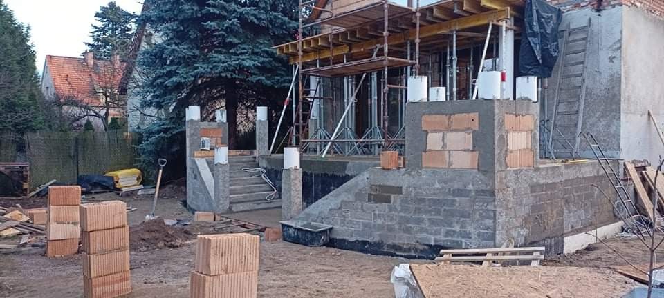 Budowa domu jednorodzinnego w trakcie: widoczne fundamenty, ściany z pustaków ceramicznych, rusztowania, przygotowane materiały budowlane i teren wokół w trakcie prac ziemnych.
